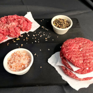 Three beef patties sitting on a black cutting board, with salt and pepper and more ground beef on wax paper