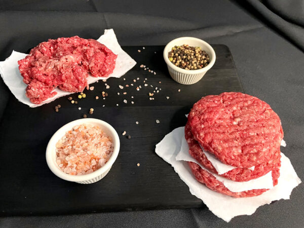 Three beef patties sitting on a black cutting board, with salt and pepper and more ground beef on wax paper