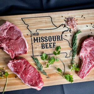 three slabs of steak on a cutting board with herbs and spices, cutting board reads Mid Missouri Beef