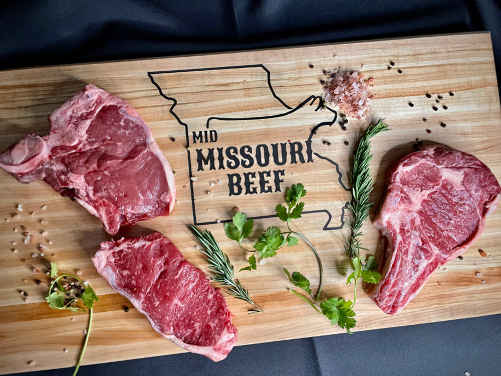 three slabs of steak on a cutting board with herbs and spices, cutting board reads Mid Missouri Beef