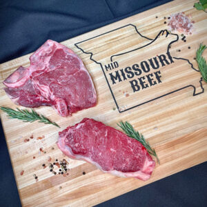 two slabs of steak on a cutting board with herbs and spices, cutting board reads Mid Missouri Beef