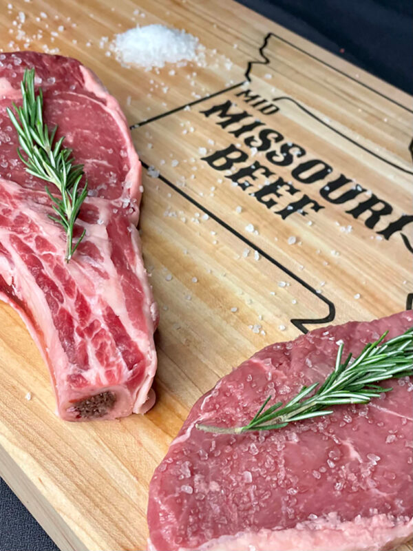 two slabs of steak on a cutting board with herbs and spices, cutting board reads Mid Missouri Beef